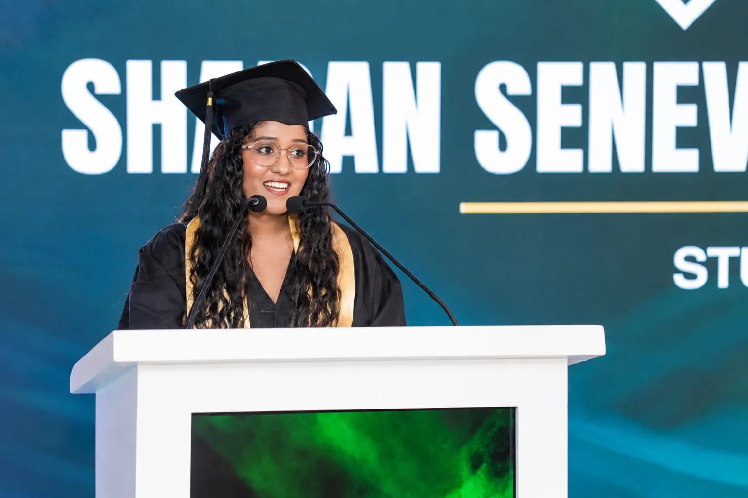 Sharan Senevirathne giving her graduation speech at the lectern, in her cap and gown